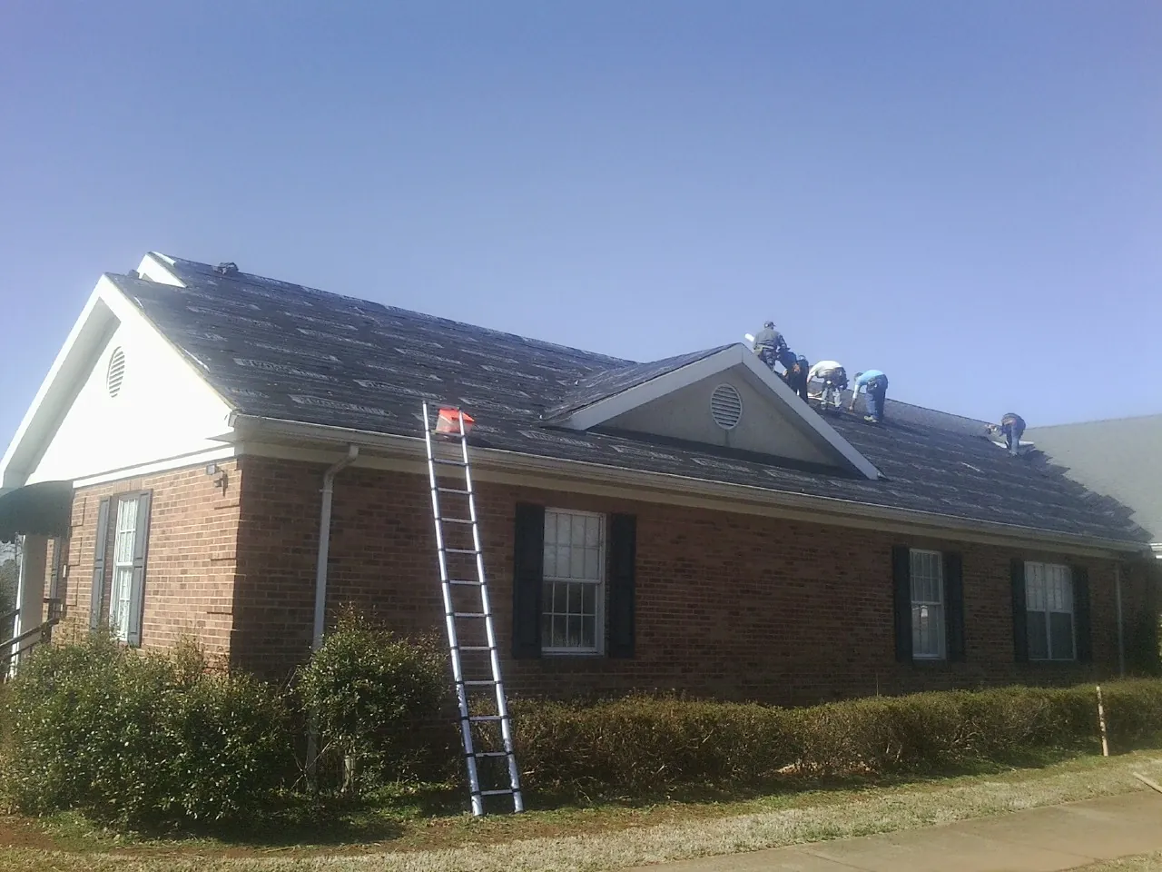 Crew tearing shingles off a brick ranch roof with a ladder set — Metal Roof Repair work by our crew in Plainview